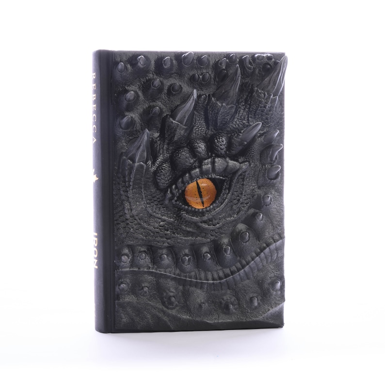 May include: Black leather-bound book with a dragon eye embossed on the cover. The eye is yellow with a black pupil. The book has the words "Rebecca Iron" printed on the spine.