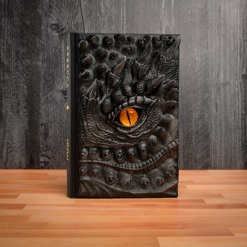 May include: A black leather-bound journal with a dragon's eye embossed on the cover. The eye is a bright orange color and the journal has the words "Rebecca Y. Iron" printed on the spine.