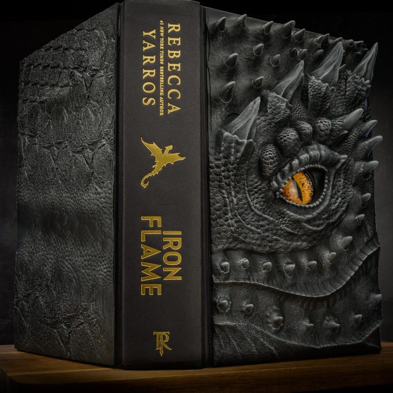 May include: A black hardcover book with a gold dragon design on the cover. The title "IRON FLAME" is written in gold lettering on the spine, along with the author's name "REBECCA YARROS".