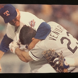 May include: A baseball player in a white and blue uniform with the letter "T" on his cap is tackling another baseball player wearing a white and grey uniform with the number "23" and the name "VENUCCI" on the back.