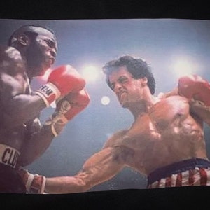 Rocky Balboa uppercutting Clubber Lang Premium T-Shirt in Mens sizes S-3XL in white or black! Printed on Kirkland brand T-Shirt!