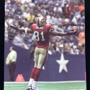 May include: A football player wearing a red and gold jersey with the number 81 and the name "OWENS" on the back. He is standing on a football pitch with a crowd of people in the background.