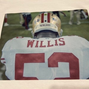 May include: A white football jersey with the number 55 and the name "WILLIS" on the back. The jersey has red trim and the player's helmet is visible in the background.