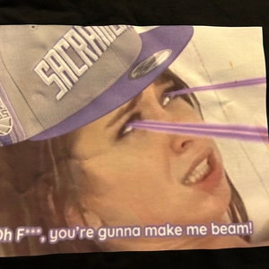 Puede incluir: Una mujer con una gorra de béisbol de los Sacramento Kings de color morado y blanco con el texto "Oh F***, you're gonna make me beam!" impreso debajo. Los ojos de la mujer brillan con rayos de luz morados.
