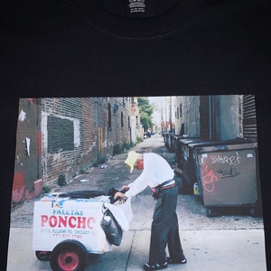 May include: A man in a yellow hat is pushing a cart with a sign that reads "Paletas Poncho" and a phone number. The cart is in an alleyway with brick walls and garbage bins.