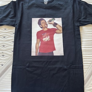 May include: Black t-shirt with a rectangular graphic of a person drinking from a bottle. The person is wearing a red shirt with a graphic design. The t-shirt is laid flat on a patterned surface.