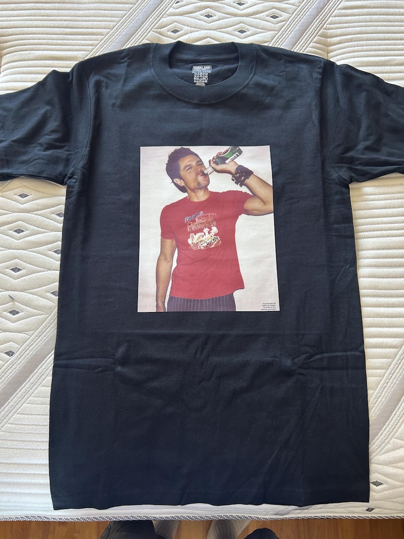 May include: Black t-shirt with a rectangular graphic of a person drinking from a bottle. The person is wearing a red shirt with a graphic design. The t-shirt is laid flat on a patterned surface.