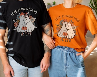 May include: Black and orange t-shirts with the text "LOVE AT HAUNT SIGHT" and a graphic of two hugging ghosts holding a pumpkin. The shirts are part of a unisex collection, also available in sweatshirt and hoodie styles.