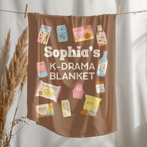 May include: A brown blanket with various food and drink illustrations, including a bottle of soju and a package of ramen. The blanket has the text "Sophia's K-DRAMA BLANKET" in white lettering.