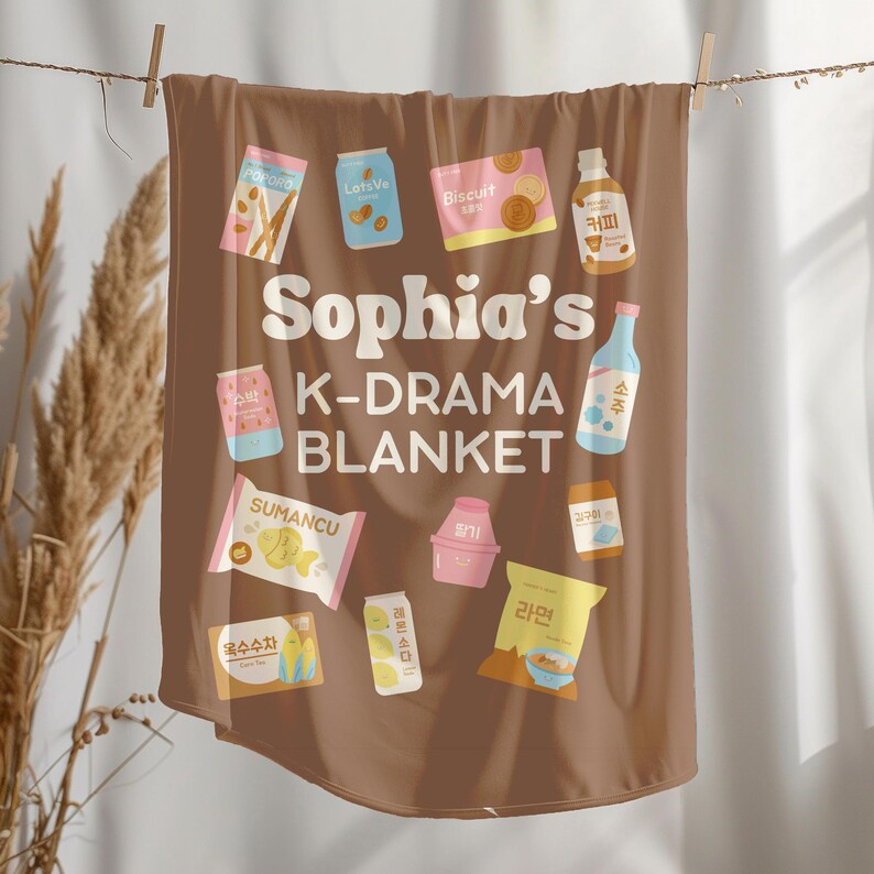 May include: A brown blanket with various food and drink illustrations, including a bottle of soju and a package of ramen. The blanket has the text "Sophia's K-DRAMA BLANKET" in white lettering.