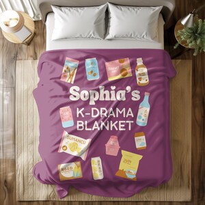 May include: A purple blanket with the text "Sophia's K-DRAMA BLANKET" and various food and drink illustrations. The blanket features images of snacks, drinks, and a bottle of soju. The blanket is on a bed with white pillows.