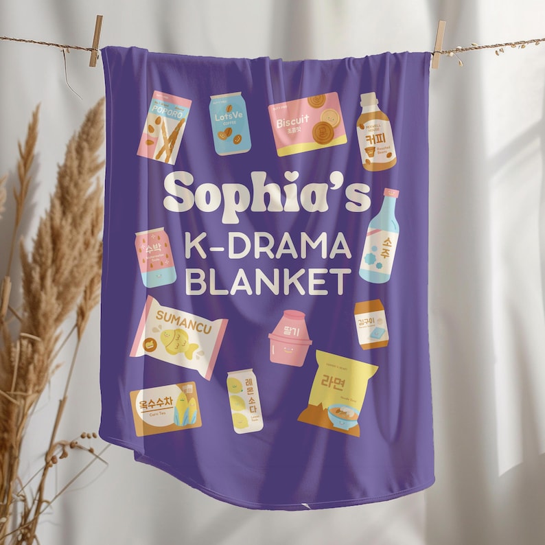 May include: A purple K-Drama blanket with the text "Sophia's K-DRAMA BLANKET". The blanket features illustrations of various Korean snacks and drinks, including biscuits, coffee, and ramen. The blanket is hanging on a clothesline.