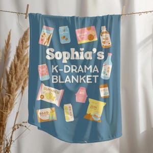 May include: A blue blanket with the text "Sophia's K-DRAMA BLANKET" and various food and drink illustrations. The blanket features images of snacks, drinks, and Korean text.