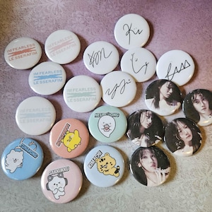 May include: A collection of round pinback buttons with various designs. Some buttons feature the text "IM FEARLESS" and "LE SSERAFIM". Other buttons have cartoon characters, signatures, and images of people. The buttons are in various colors.