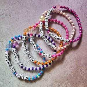 May include: Five beaded bracelets in various colors, including pink, purple, blue, and orange. Each bracelet features white letter beads spelling out names like "Rainbow Dash" and "Sparkle". The bracelets are on a light purple surface.