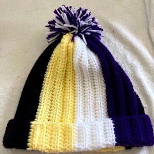 Könnte beinhalten: Eine gestrickte Mütze mit schwarzen, gelben, weißen und violetten Streifen und einem weißen und violetten Bommel oben.