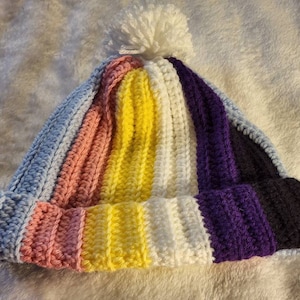 Op de afbeelding: Een gebreide beanie met een witte pompon bovenop. De muts is gestreept met lichtblauw, roze, geel, wit, paars en zwart.