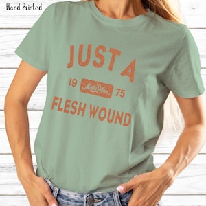 May include: Sage green t-shirt with the text "JUST A FLESH WOUND" in orange. The text includes the year "1975" and the words "Monty Python" in a rectangular box. The words "Hand Painted" are in the upper left corner.