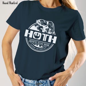 May include: Navy blue t-shirt with a white graphic featuring the word "HOTH" and the phrases "DEFEND ECHO BASE" and "SUPPORT THE REBELLION". The graphic includes an image of a vehicle and a planet. The words "Hand Painted" are in the top left corner.