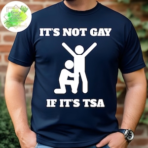 May include: Navy blue t-shirt with the text "IT'S NOT GAY IF IT'S TSA" in white block letters. The shirt features a white graphic of two stick figures. A Fern's Craft Corner logo is in the upper left corner.