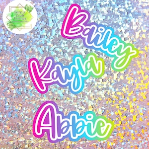 May include: Three custom name stickers: "Bailey", "Kayla", and "Abbie", in a vibrant gradient of pink, blue, and green, outlined in white. The background is a shimmering, iridescent surface. A logo in a gold frame with the text "Fern's Craft Corner Handmade Gifts" is in the upper left corner.