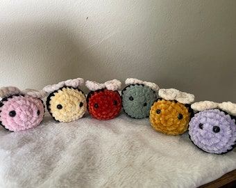 May include: Seven crocheted bee plushies in different colors, including pink, yellow, red, gray, gold, and purple. Each bee has black eyes and a black stripe around the middle.