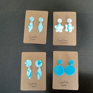 May include: Four pairs of handmade polymer clay earrings on brown card stock. The earrings are in various shapes and colors, including blue, white, and turquoise. The card stock has the text "Creation Cloco" written on it.
