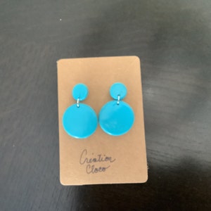 May include: A pair of turquoise blue dangle earrings with a smaller circle on top of a larger circle. The earrings are on a brown card with the text "Creation Cloco" written on it.