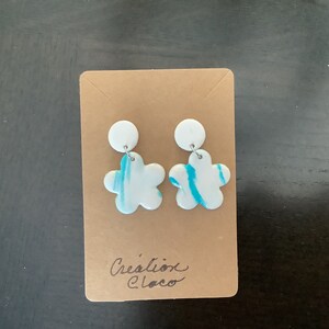May include: A pair of white flower-shaped earrings with blue swirl designs. The earrings are displayed on a brown card with the text "Cr&eacute;ation Cloco".