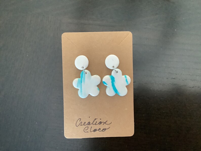 May include: A pair of white flower-shaped earrings with blue swirl designs. The earrings are displayed on a brown card with the text "Cr&eacute;ation Cloco".