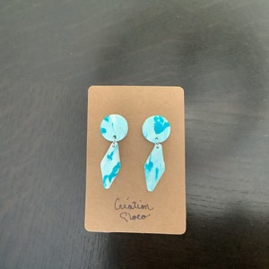 May include: A pair of blue and white patterned earrings with a diamond shape dangle. The earrings are displayed on a brown card with the text "Cr&eacute;ation Floco".