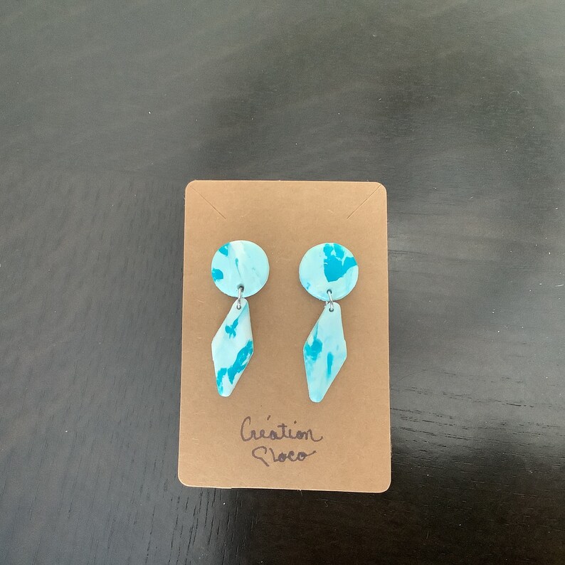 May include: A pair of blue and white patterned earrings with a diamond shape dangle. The earrings are displayed on a brown card with the text "Cr&eacute;ation Floco".