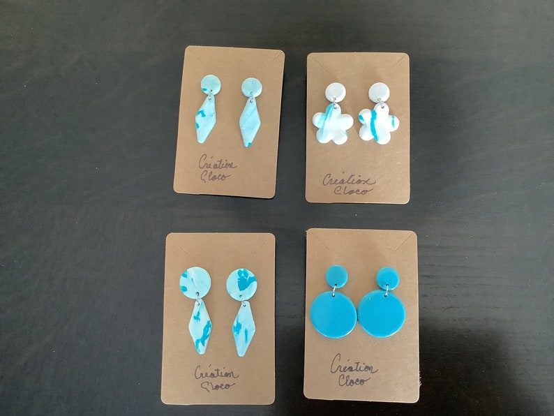 May include: Four pairs of handmade polymer clay earrings on brown card stock. The earrings are in various shapes and colors, including blue, white, and turquoise. The card stock has the text "Creation Cloco" written on it.