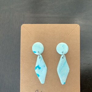 May include: A pair of light blue and turquoise earrings with a diamond shape and a small round accent. The earrings are on a brown card with the text "Creation Blocco".