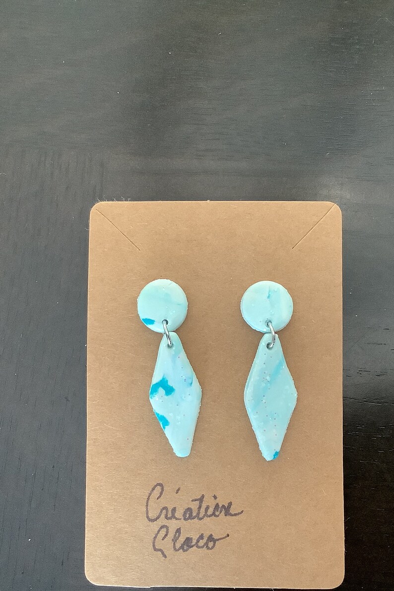 May include: A pair of light blue and turquoise earrings with a diamond shape and a small round accent. The earrings are on a brown card with the text "Creation Blocco".