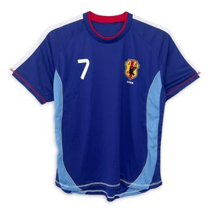 Può includere: Una maglia da calcio blu con dettagli bianchi e blu chiaro. La maglia ha un colletto rosso e bordi rossi sulle maniche. Il numero "7" è stampato sul davanti della maglia. La maglia ha anche un logo con il testo "JFA" e "GIAPPONE" stampato sul davanti.