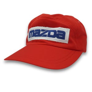 Könnte beinhalten: Rote Baseballkappe mit gebogenem Schirm. Auf der Kappe steht das Wort "MAZDA" in blauen Blockbuchstaben auf einem weißen rechteckigen Aufnäher. Die Kappe ist aus leichtem Material und für Outdoor-Aktivitäten geeignet.