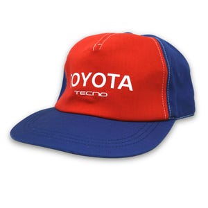 Peut inclure: Casquette de baseball rouge et bleue avec le mot "TOYOTA" en blanc. La casquette a une visière bleue et une couronne rouge. Le mot "TECNO" est imprimé en dessous de "TOYOTA".