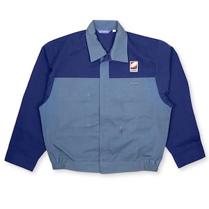 May include: A two-tone work jacket in navy blue and light blue. The jacket features a collared neckline, long sleeves, and a ribbed hem. A small logo is visible on the chest. The jacket has a button-up front and two front pockets.