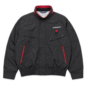 May include: A black jacket featuring a high collar with red trim, a front zipper closure, and a chest pocket with a red loop and the word NISSAN printed in white. The garment includes two side pockets with red accents and an elasticized hem.