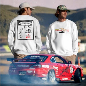 May include: White sweatshirts with graphic designs and a red sports car. One sweatshirt features a "Caution Wide Turns" sign. The other sweatshirt has the text "It's Time For Action!" The red car has racing decals and is drifting on a track.