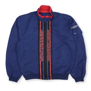 Op de afbeelding: Een marineblauwe bomberjack met een rode kraag en manchetten. De jas heeft verticale zwarte, rode en blauwe strepen over de voorkant met de tekst "NISSEKI GAS" in het rood. Een klein zakje zit op de linkermouw.