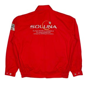 Puede incluir: Chaqueta roja brillante con la palabra "SOLUNA" bordada en blanco en la espalda. El texto "The Project SOLUNA is a crystallization of new standard of family sedan developed by TOYOTA for 21st century" está debajo del logotipo. Un pequeño parche está en la manga izquierda.