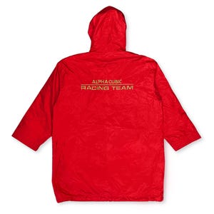 Könnte beinhalten: Eine leuchtend rote Kapuzenjacke mit kurzen Ärmeln. Auf der Rückseite der Jacke steht in goldener Schrift "ALPHA CUBIC RACING TEAM". Die Jacke besteht aus einem leichten, möglicherweise wasserabweisenden Material.