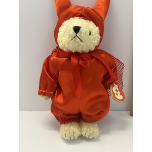 Puede incluir: Un oso de peluche color crema que viste un disfraz de diablo de satén naranja brillante con una capucha que presenta dos cuernos puntiagudos. El conjunto incluye un lazo de cinta naranja a juego en el cuello y una capa de malla roja, con una etiqueta Ty en forma de corazón.