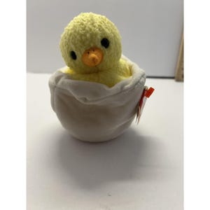 May include: A plush yellow chick with black eyes and an orange beak sits inside a soft, off-white egg. The chick is made of a fuzzy material. A red tag is attached to the egg. The toy is designed to look like a baby chick hatching from its egg.