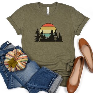 K&ouml;nnte beinhalten: Ein gr&uuml;nes T-Shirt mit einem Retro-Sonnenuntergang-Motiv mit einem Disc-Golf-Korb und B&auml;umen. Das Motiv ist in Orange-, Gelb- und Blaut&ouml;nen gehalten.
