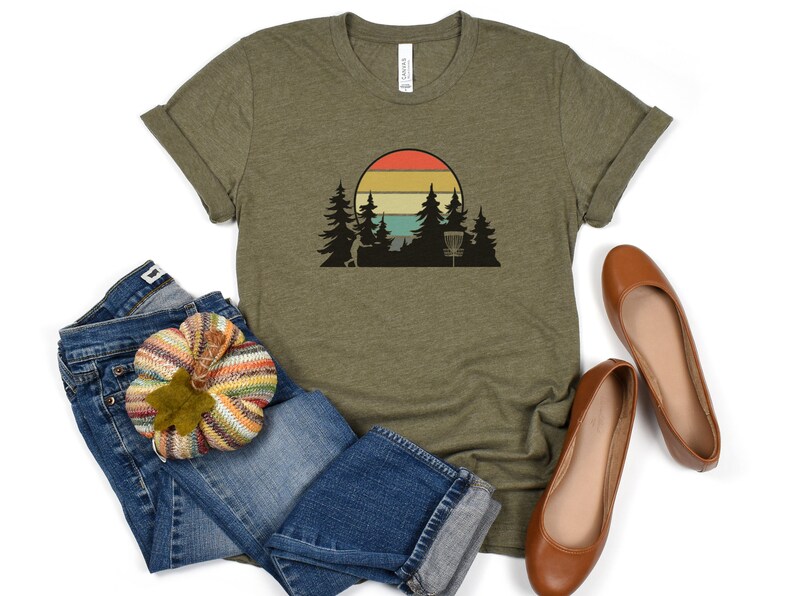 K&ouml;nnte beinhalten: Ein gr&uuml;nes T-Shirt mit einem Retro-Sonnenuntergang-Motiv mit einem Disc-Golf-Korb und B&auml;umen. Das Motiv ist in Orange-, Gelb- und Blaut&ouml;nen gehalten.