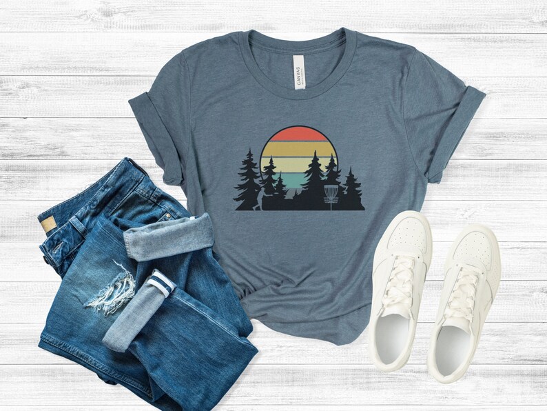 K&ouml;nnte beinhalten: Ein blaues T-Shirt mit einem Retro-Sonnenuntergang-Motiv mit einem Disc-Golf-Korb und B&auml;umen. Das Motiv ist in Orange-, Gelb-, Gr&uuml;n- und Blaut&ouml;nen gehalten.