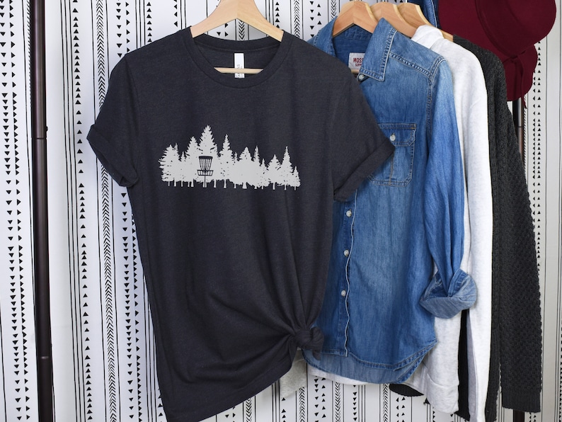 Puede incluir: Una camiseta gris oscuro con una silueta blanca de una cesta de disco golf y &aacute;rboles. La camiseta est&aacute; colgada en una percha con otras prendas.